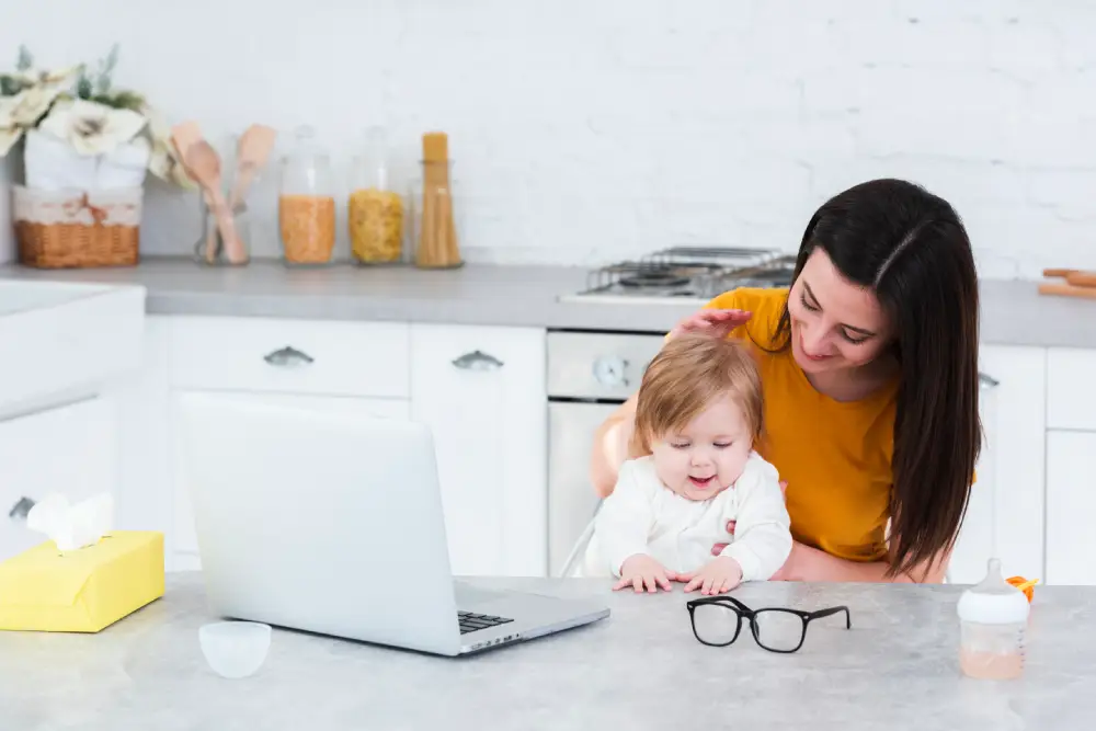 Woman holding a baby in the kitchen while using a laptop